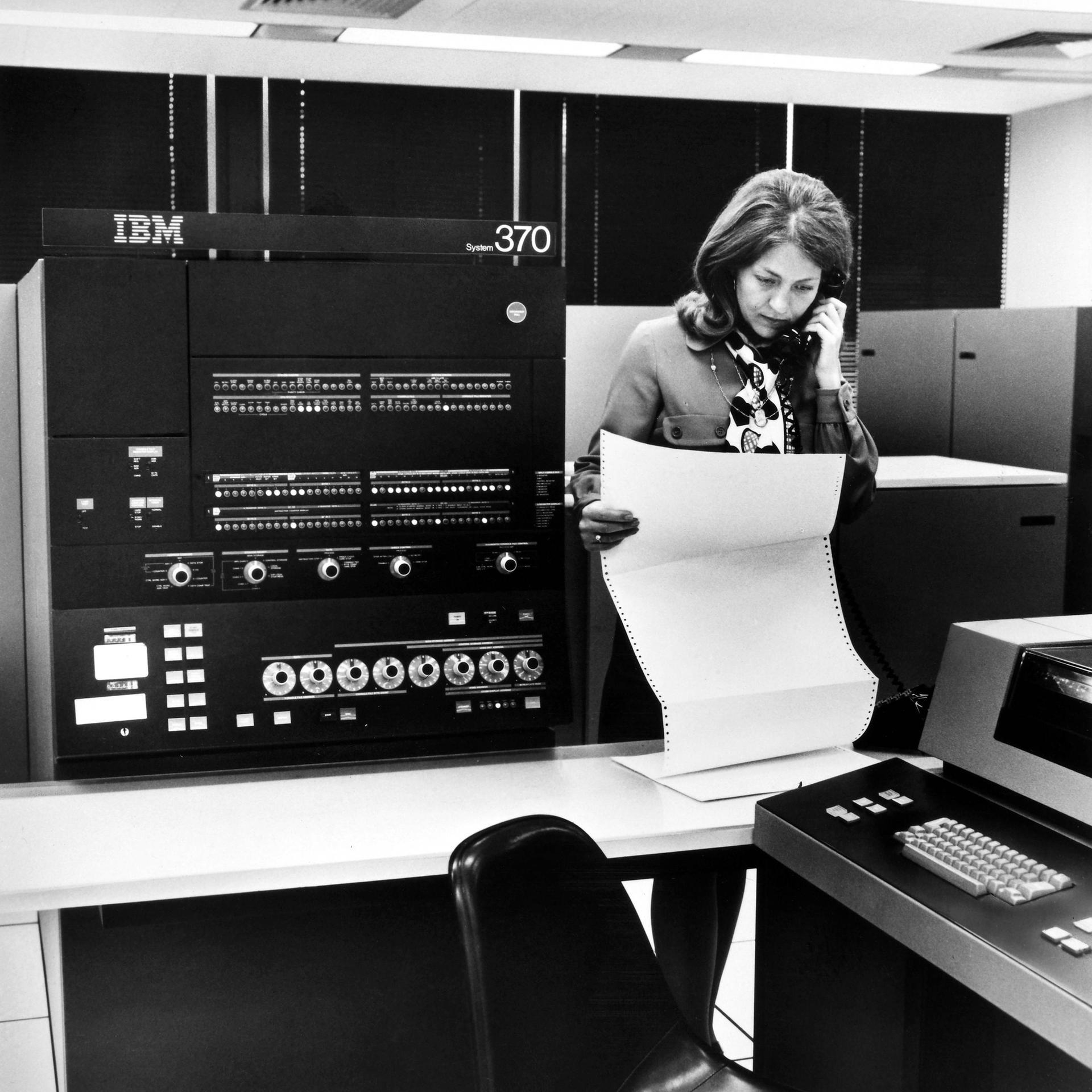 Schwarzweißfoto eines IBM 370 Großrechners. Davor eine Frau mit einem Endlospapierausdruck. Außerdem telefoniert sie noch.