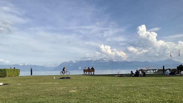 Vue du Léman. Cycliste, personnes sur un banc. Dans l’arrière-plan, les Alpes et des nuages.