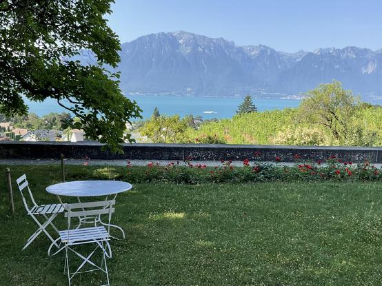 View from the Domaine de la Doges (La Tour-de-Peilz) over the Léman. In the foreground a small white bistro table with two chairs, in the background the Alps.
