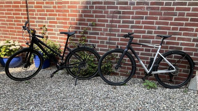 Two bikes standing in front of a brick wall: on the left, a matte black Grecos Urban Belt with a trapezoid frame); on the right, a black and silver Scott Metrix 20 EQ.