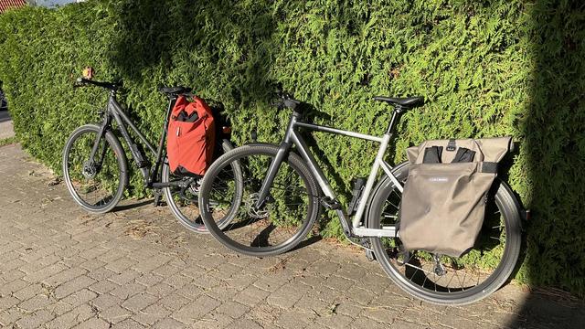 Two bikes standing in front of a hedge: on the left, a matte black Grecos Urban Belt with an Ortlieb Vario Lite backpack/bag; on the right, a black and silver Scott Metrix 20 EQ with a Vario 26L.